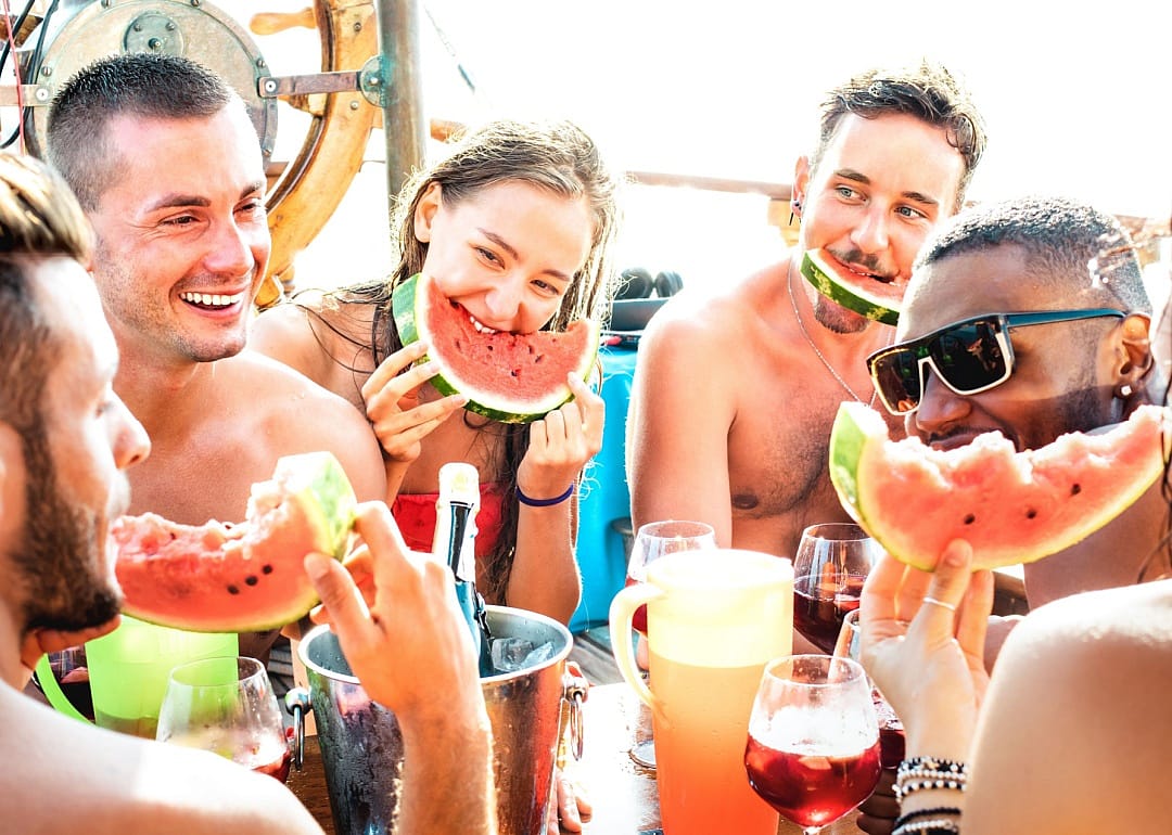 A group of young friends smiling while eating fresh watermelon slices outdoors. 