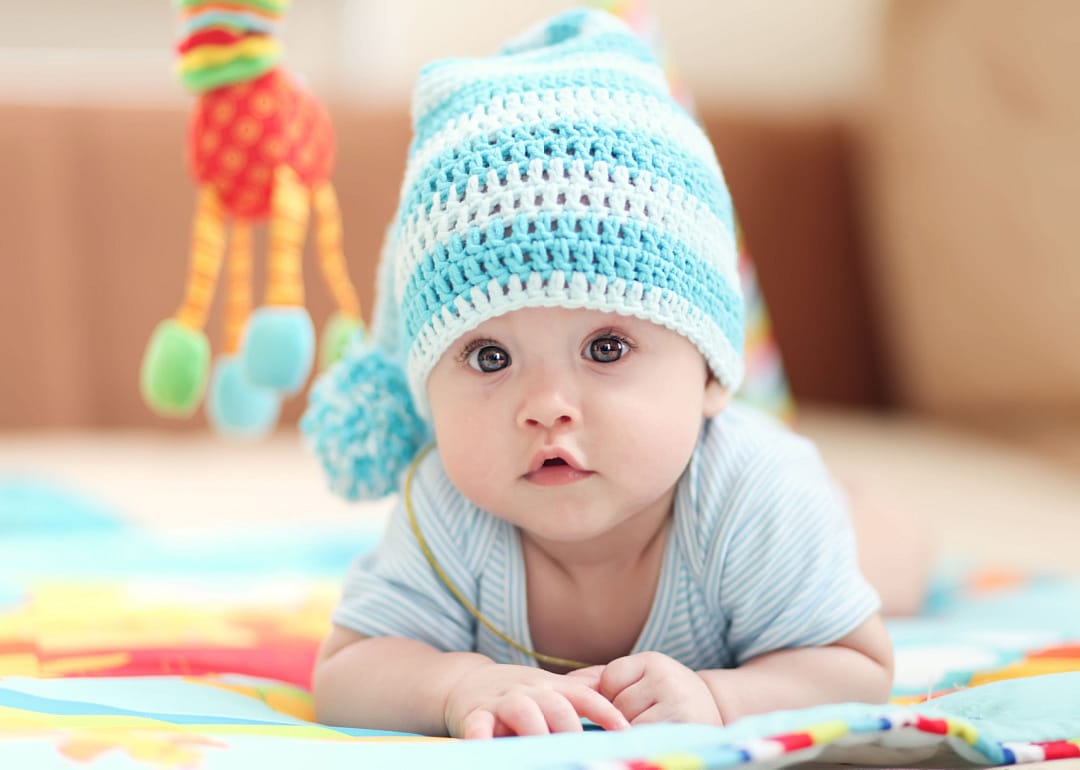 Baby girl lying on a children's rug in the white-blue cap.