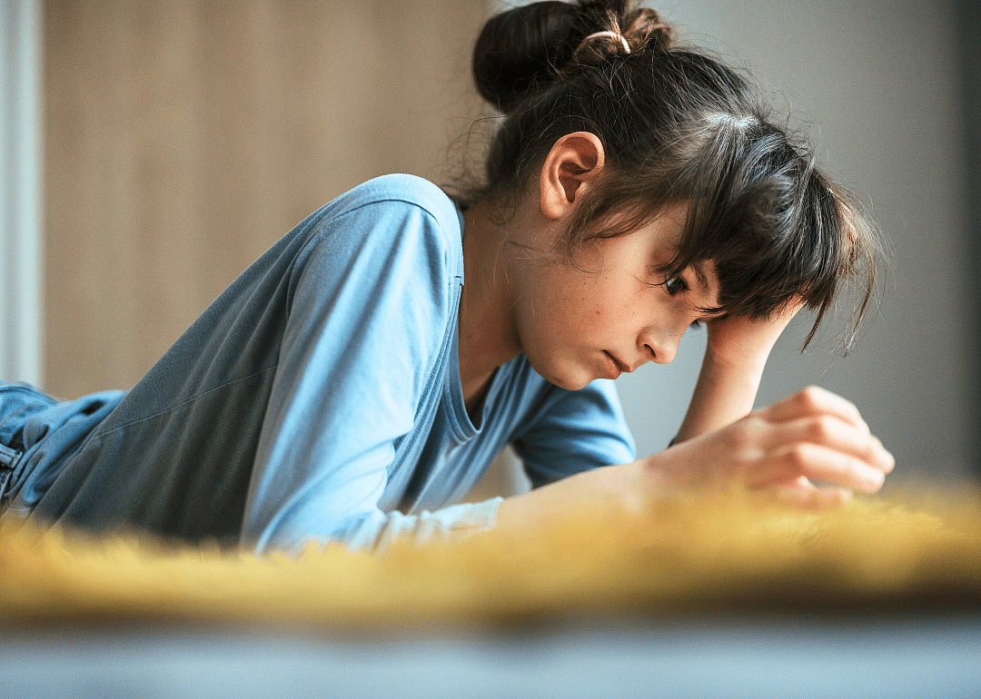 A young girl lying on her stomach. She is resting her head on one hand while looking down at something in front of her.