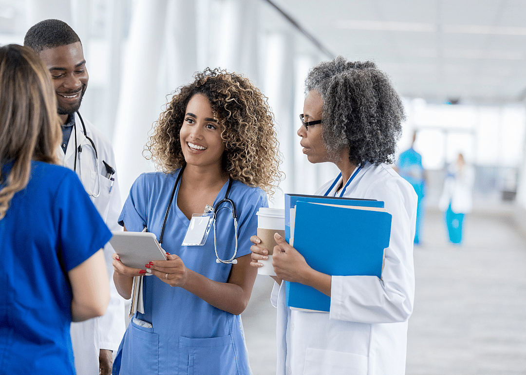 A group of medical staff, three women and a man standing close.  A young woman holding a flat device in her hands looking at the woman in front of her.  