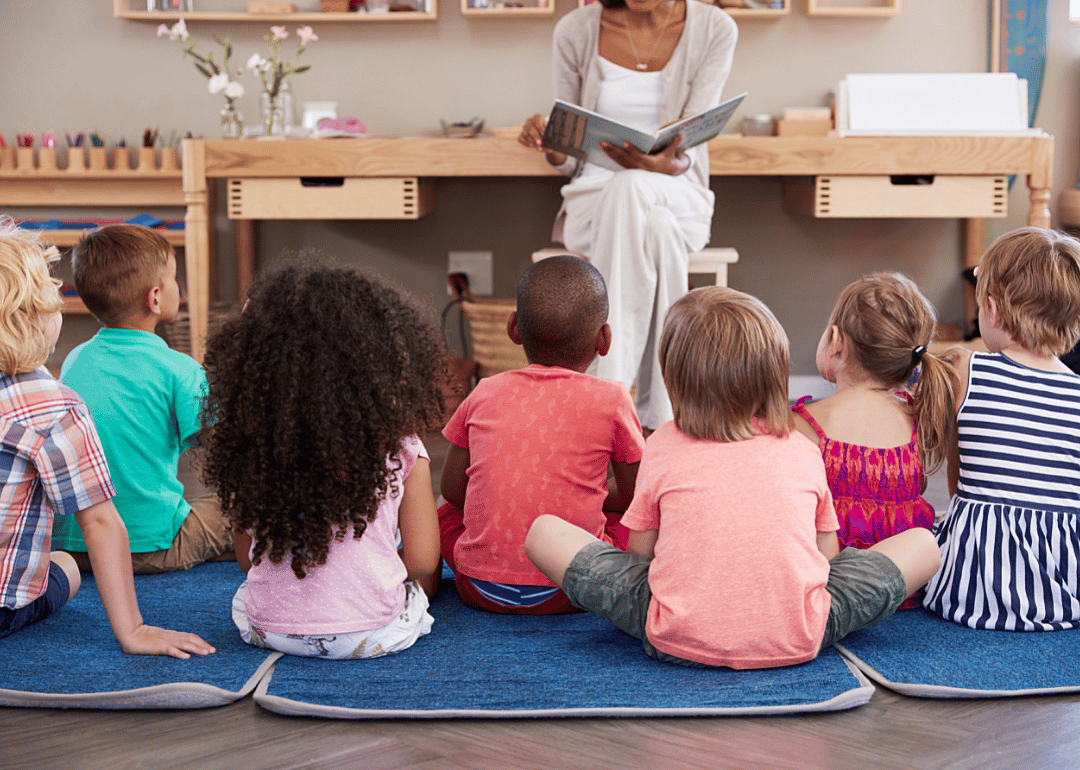 Young children sat on floor mats listening to their teacher read from a book.