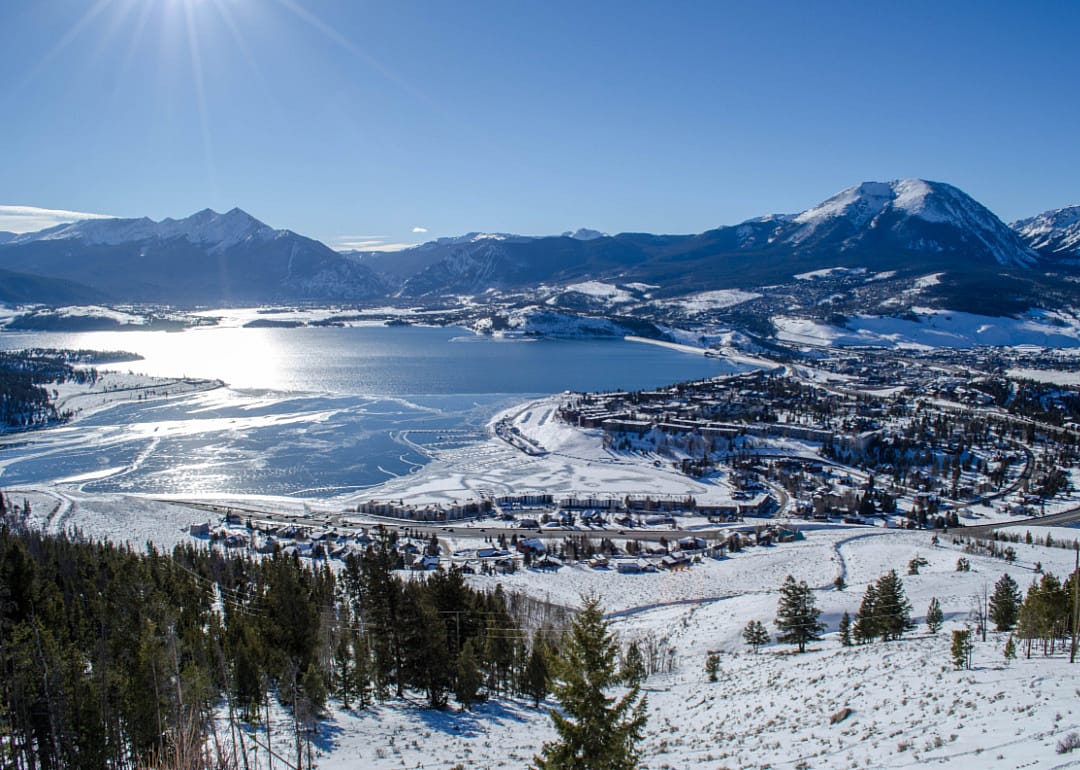 An aerial view of a vast partially frozen lake and town surrounded by tall snowy mountains.