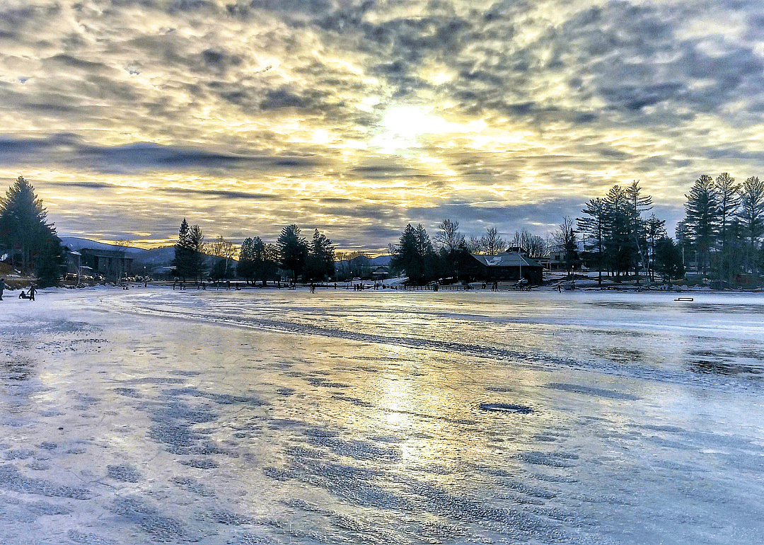 People ice skating on a frozen lake surrounded by tall trees and mountains in the background.