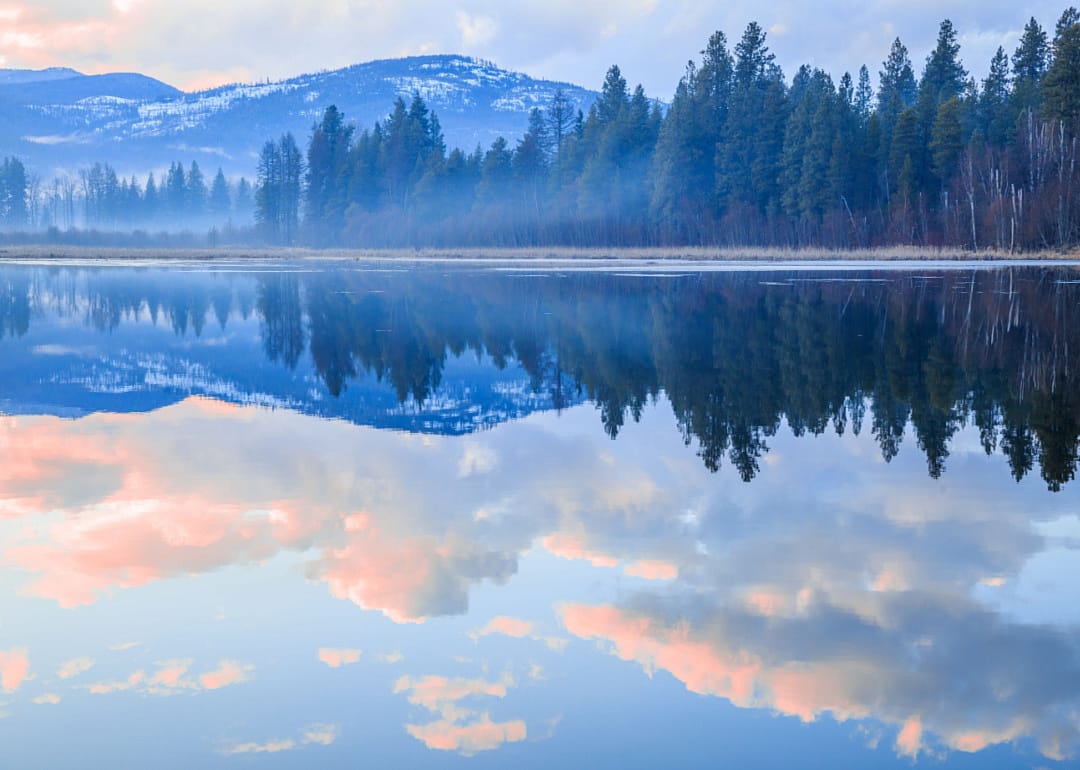 A lake with tall trees at the sunset with the reflection of the mountains and clouds in the water.