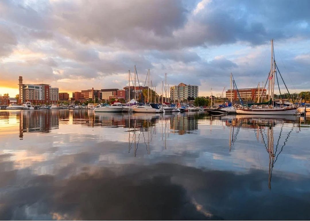 Sunset over a harbor filled with sailboats. The city skyline is visible in the distance.