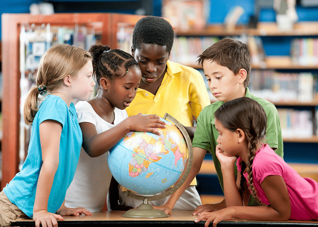 A group of five young children gathered around a globe in a classroom or library setting. The children, who are of different ethnic backgrounds, are all focused on the globe.