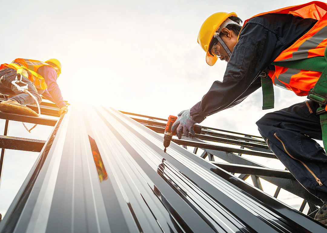 Two men wearing hard hats and orange vests climbing a metal structure. One is drilling into a huge metal sheet; the other holds the metal sheet for him. 