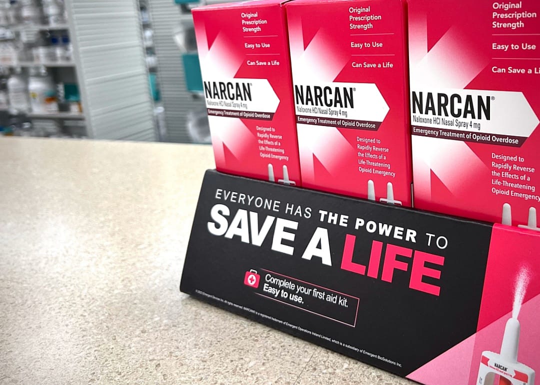 A close up of three boxes of Narcan medication on the counter at a pharmacy with a sign saying, everyone has a power to save a life.
