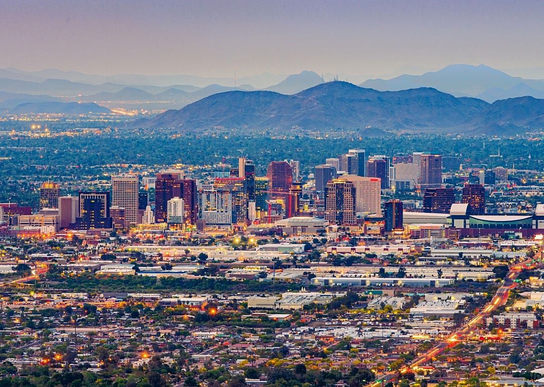 A city view at dusk, with the urban lights twinkling. A range of mountains in the background. The city has a mix of high-rise buildings concentrated in the center, with lower development spreading outwards.