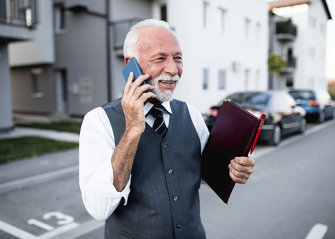 An older male real estate agent on a phone holding a leather folder.