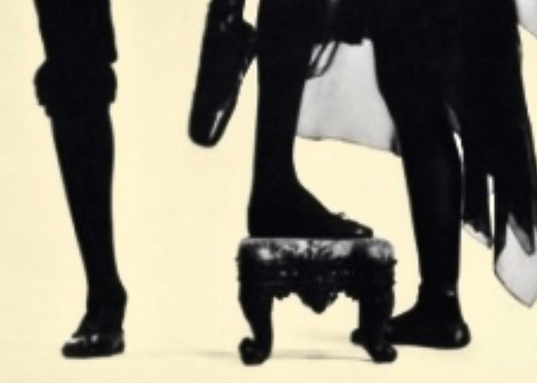 Two pairs of feet in black shoes, a small stool against a beige background.