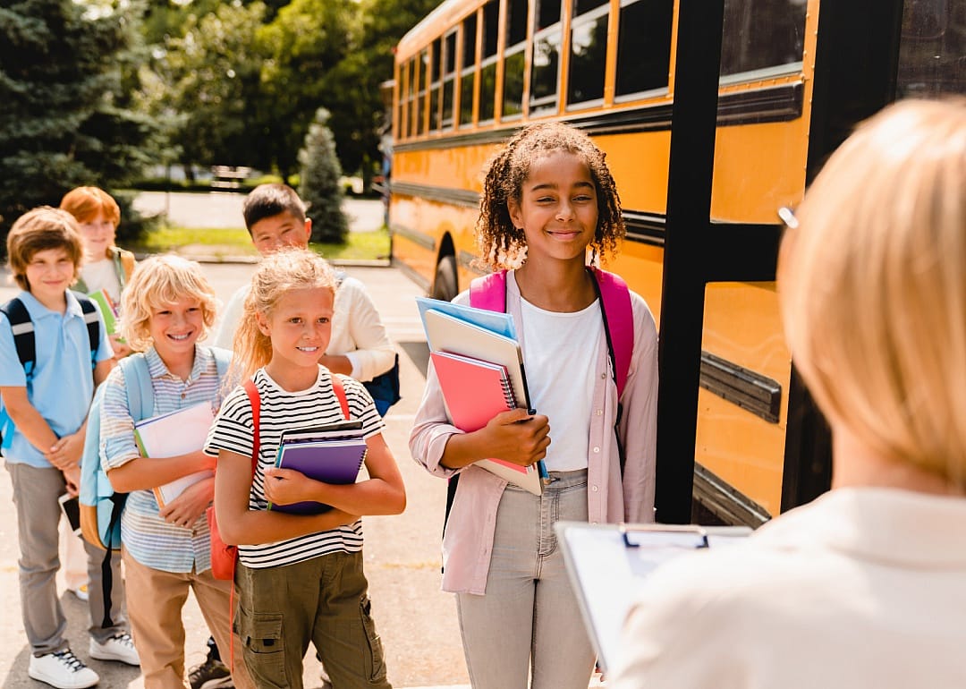 A line of schoolkids with backpacks some holding notebooks standing by schoolbus.