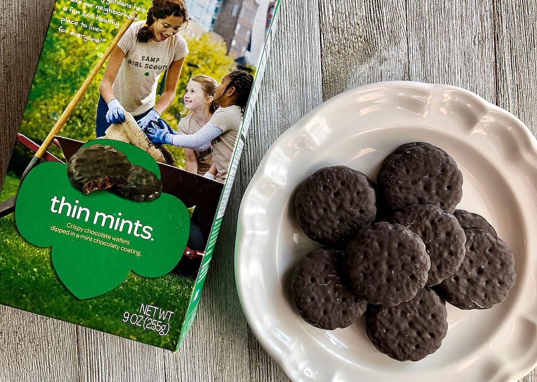 Box of Girl Scout Thin Mints with a few on a white plate on a wooden surface.