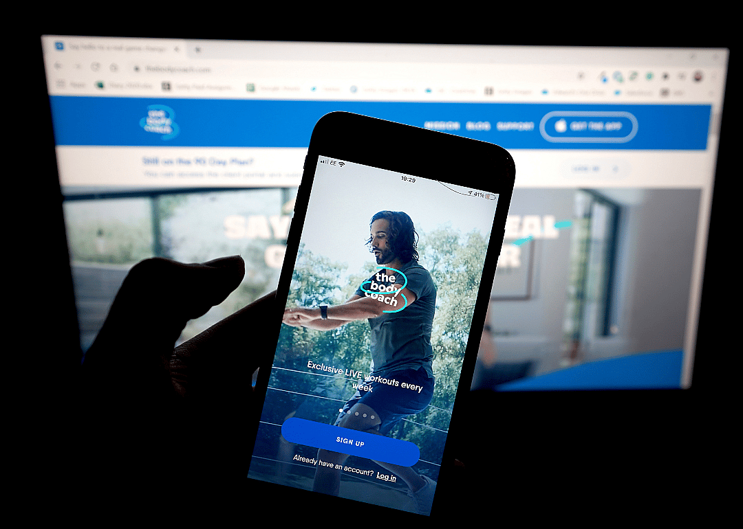 A person holding a phone showing a workout app in front of a computer.
