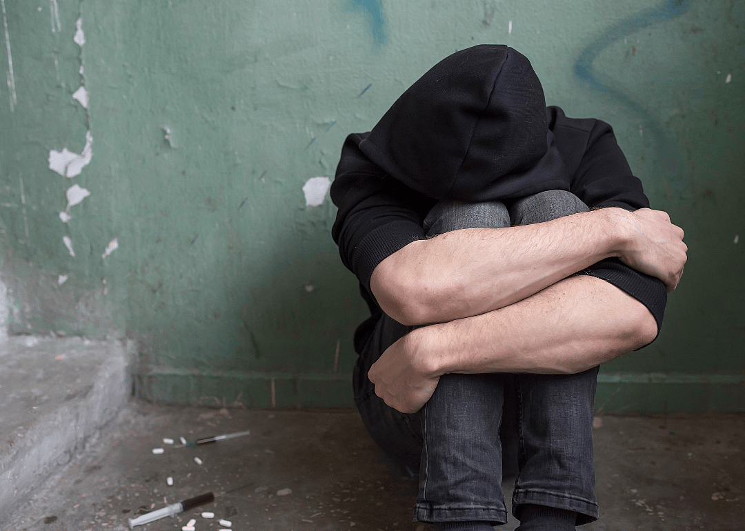 A teenager with their head resting on their knees sits on the ground against a deteriorating wall, with pills and syringes on the ground next to them.