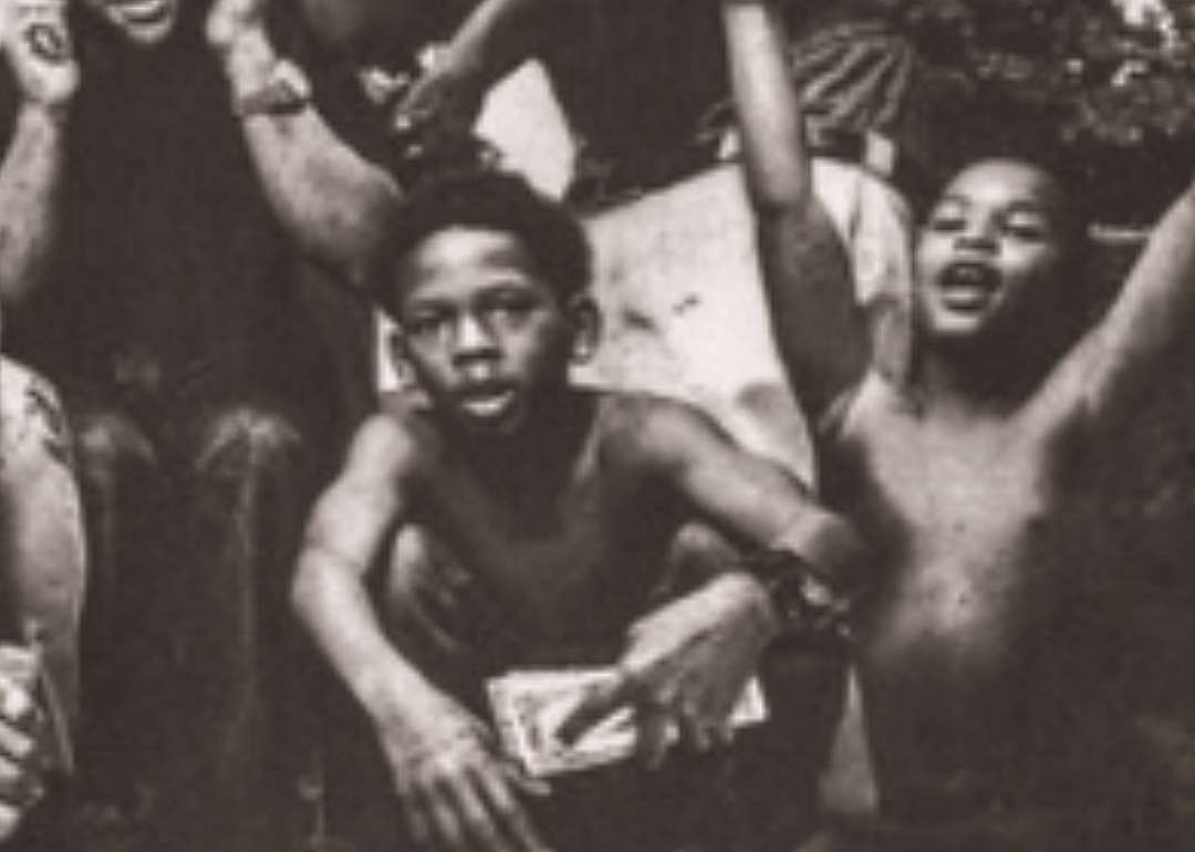 Black-and-white photo of two small shirtless Black boys who are part of a larger crowd.