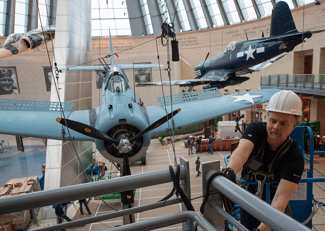 Lance Retallick, a technician with Weiss Theatrical Solutions, securing a safety line as a World War II SBD Dauntless dive bomber is craned into position at the National Museum of the Marine Corps in Triangle