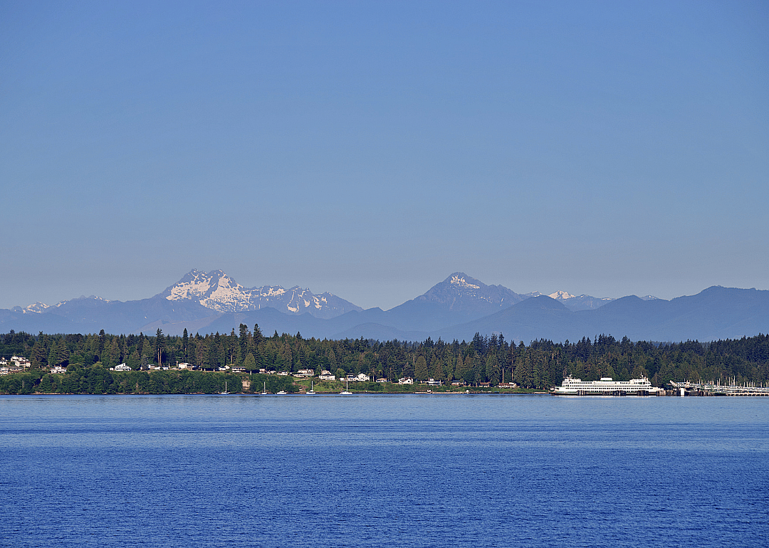 A town situated by the body of water with tall snowy mountain peaks and green trees in the background.