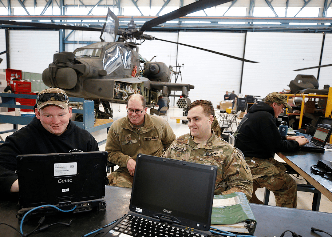 Military personnel on computers in front of a helicopter in a bright warehouse.