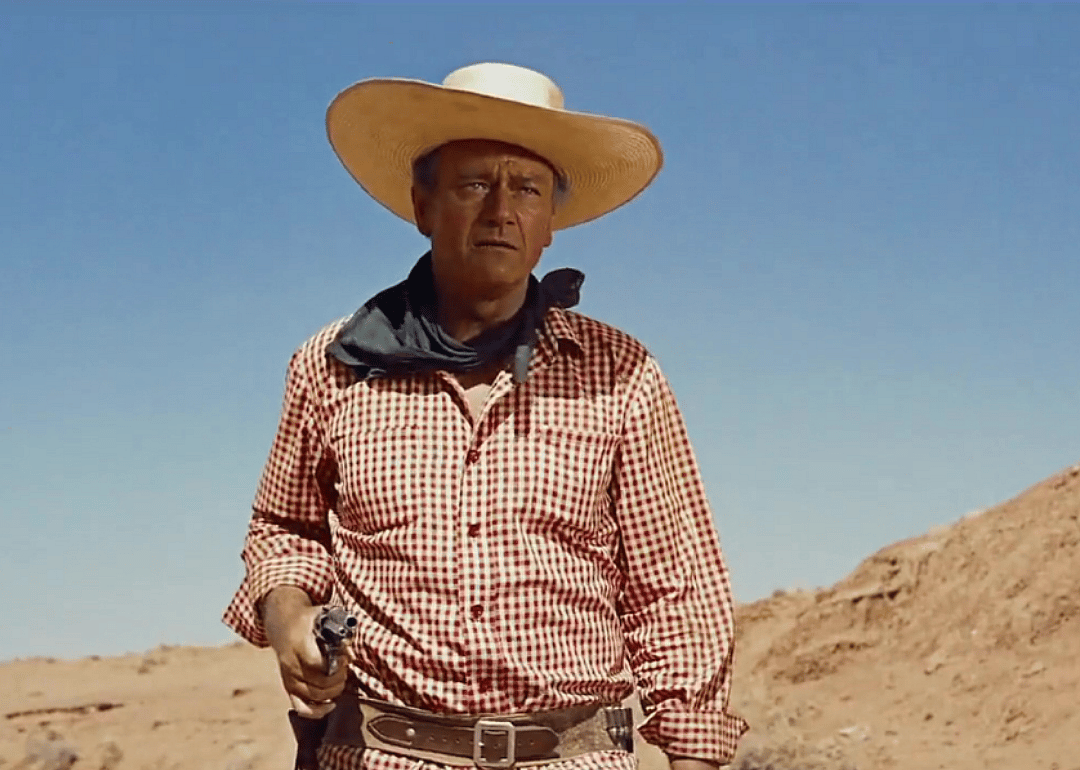 John Wayne, wearing a red and white plaid shirt, cowboy hat and dark bandana around his neck, holds a pistol.