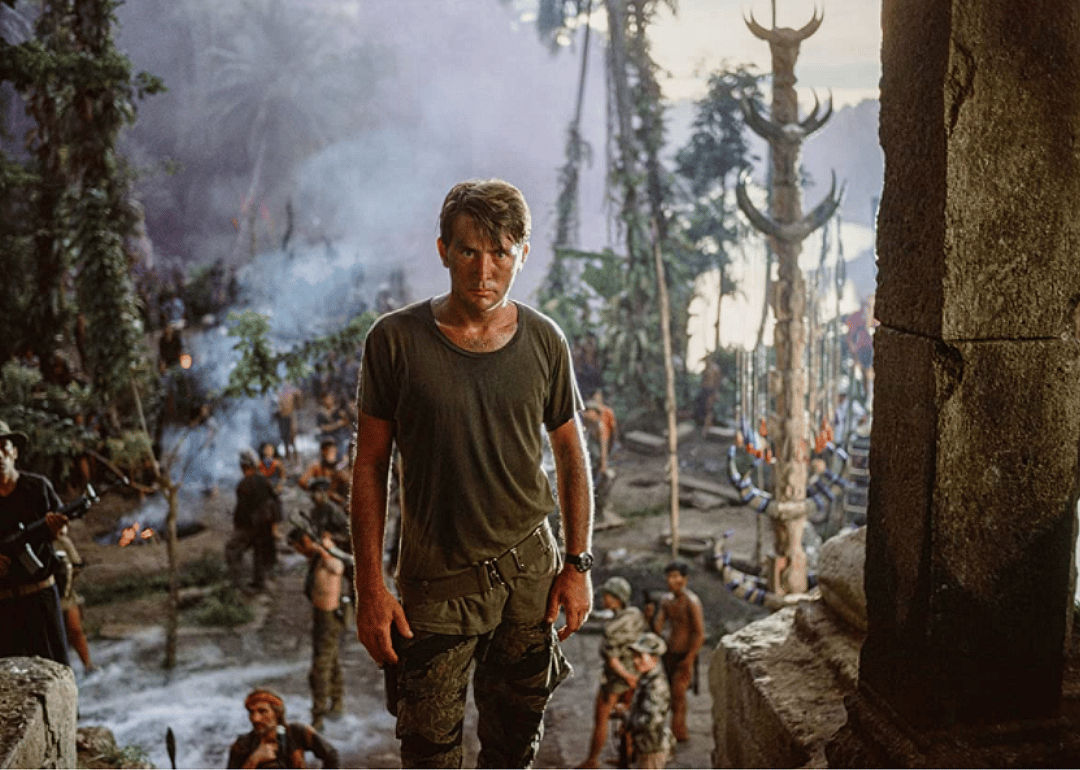 Martin Sheen in camo pants and a dark green shirt at the top of stairs, surrounded by soldiers.