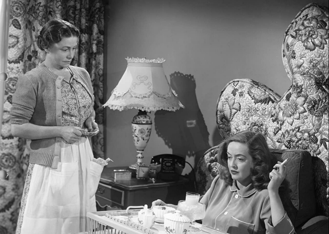 A woman smoking in bed looking angry while an older woman stands at her bedside.