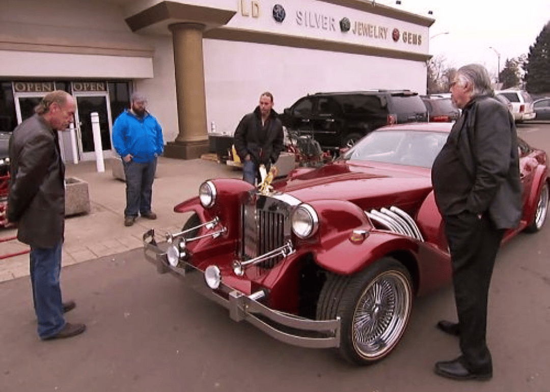 Men walk around a classic car in front of a pawn shop.