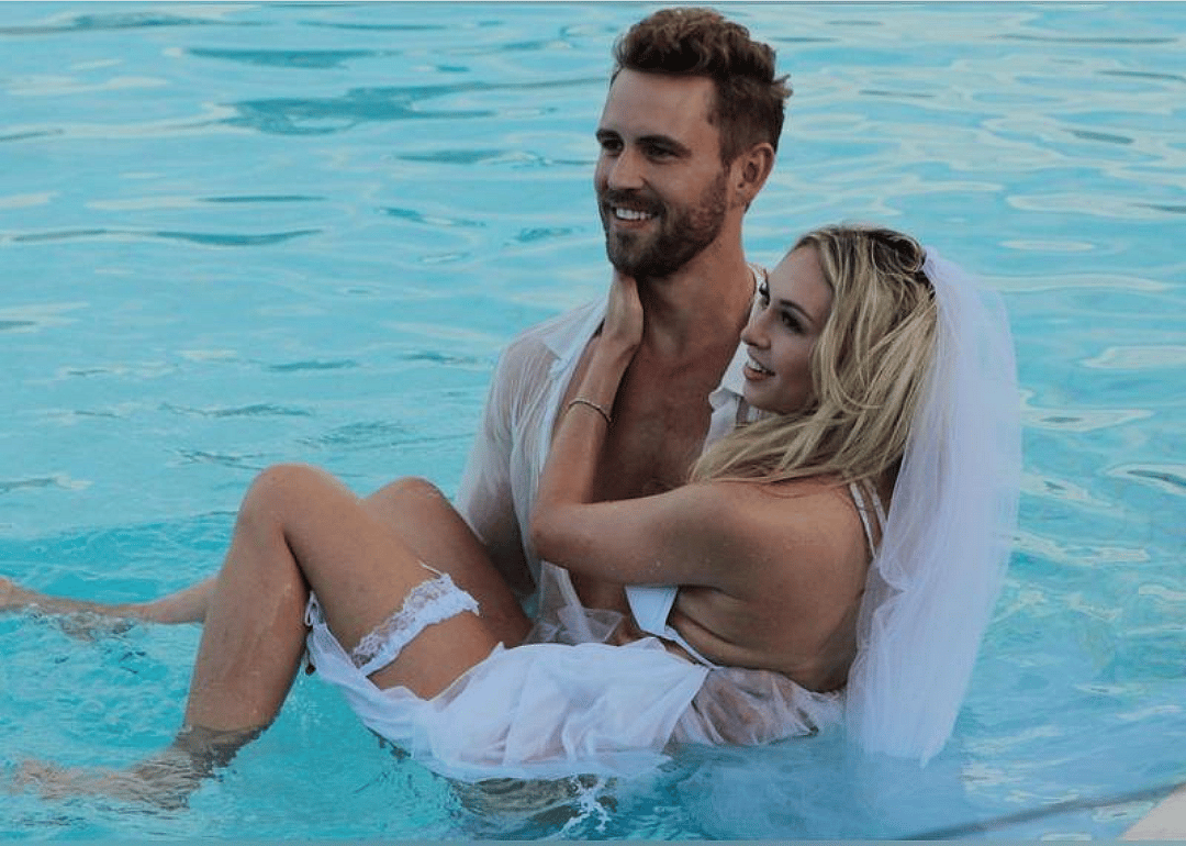 A man in a white shirt stands in a pool holding a woman in a wedding dress.
