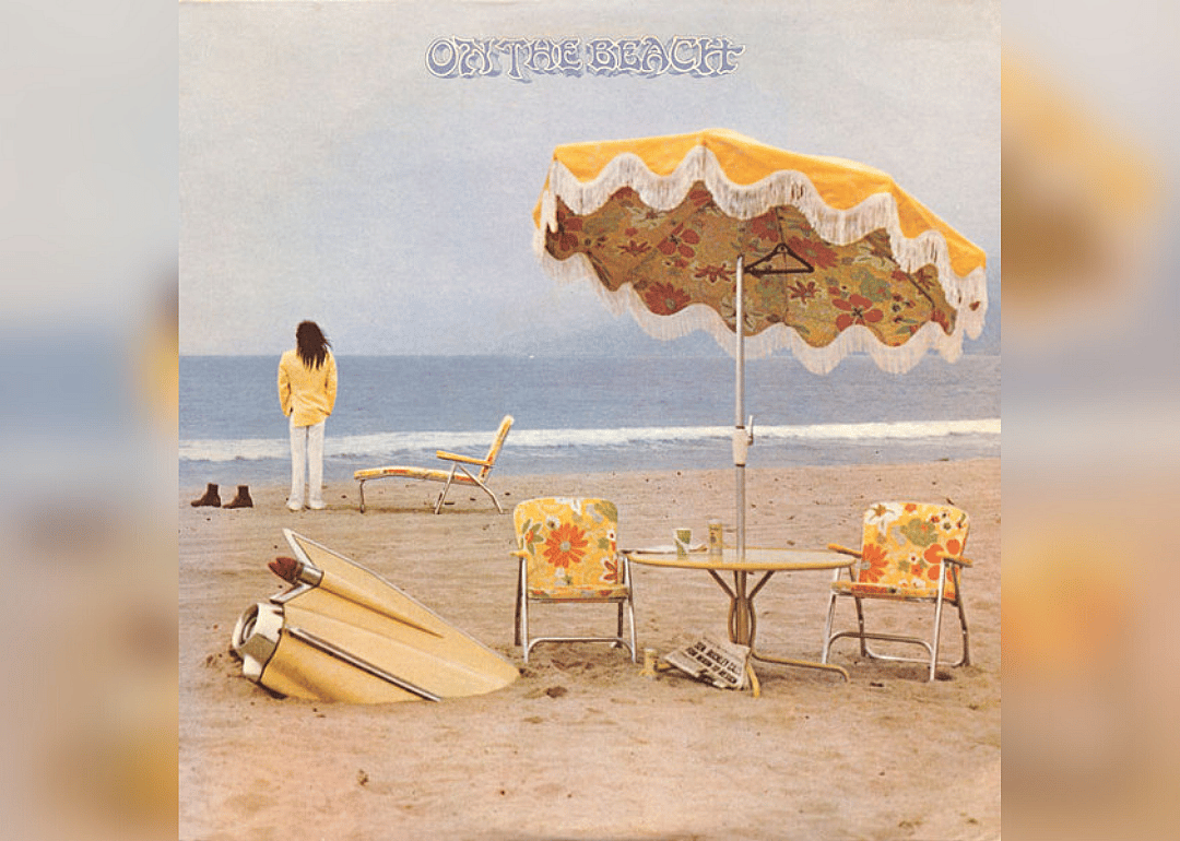 Neil Young standing on the beach in a yellow blazer looking at the ocean and everything on the beach is also yellow, including a table and chairs with umbrella..