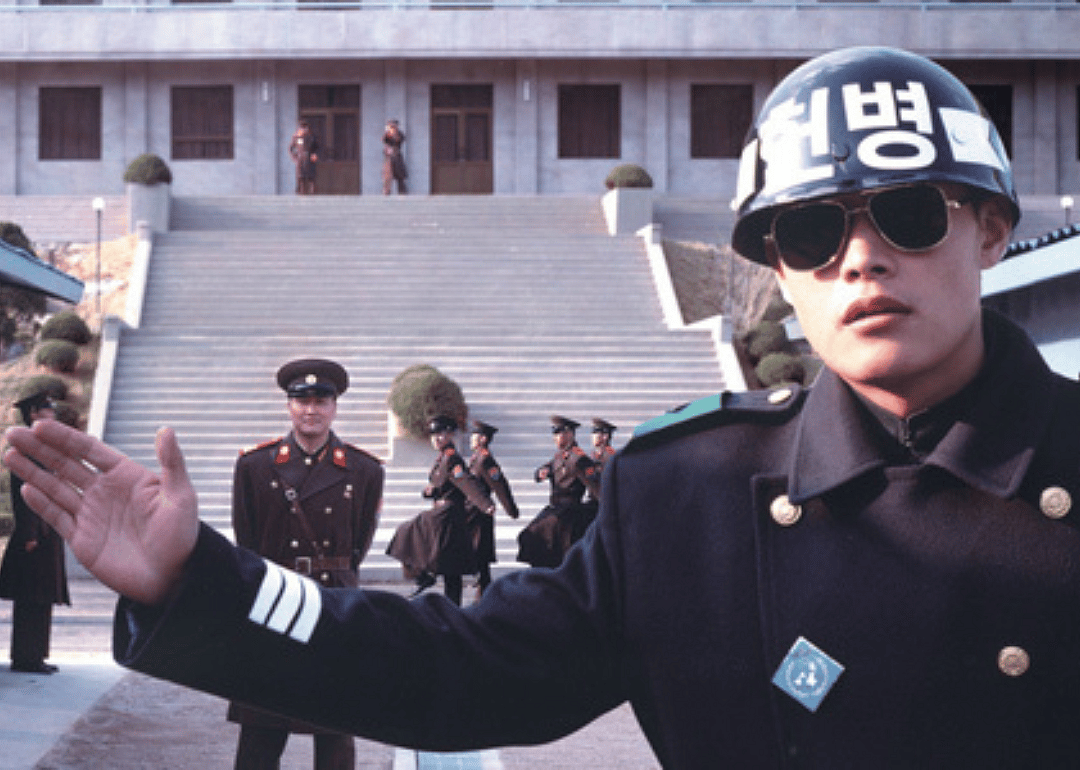 A man in uniform at a border crossing holds his hand out to stop with guards behind him.