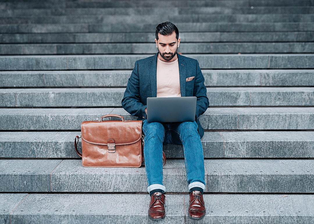 A man with a leather briefcase sitting on steps while working on his laptop.