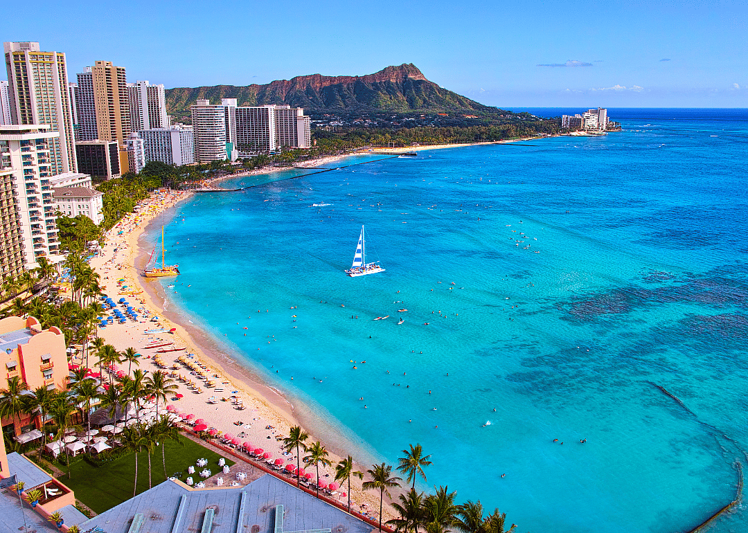 Boats on turquoise water and umbrellas on a crowded beach next to tall buildings.