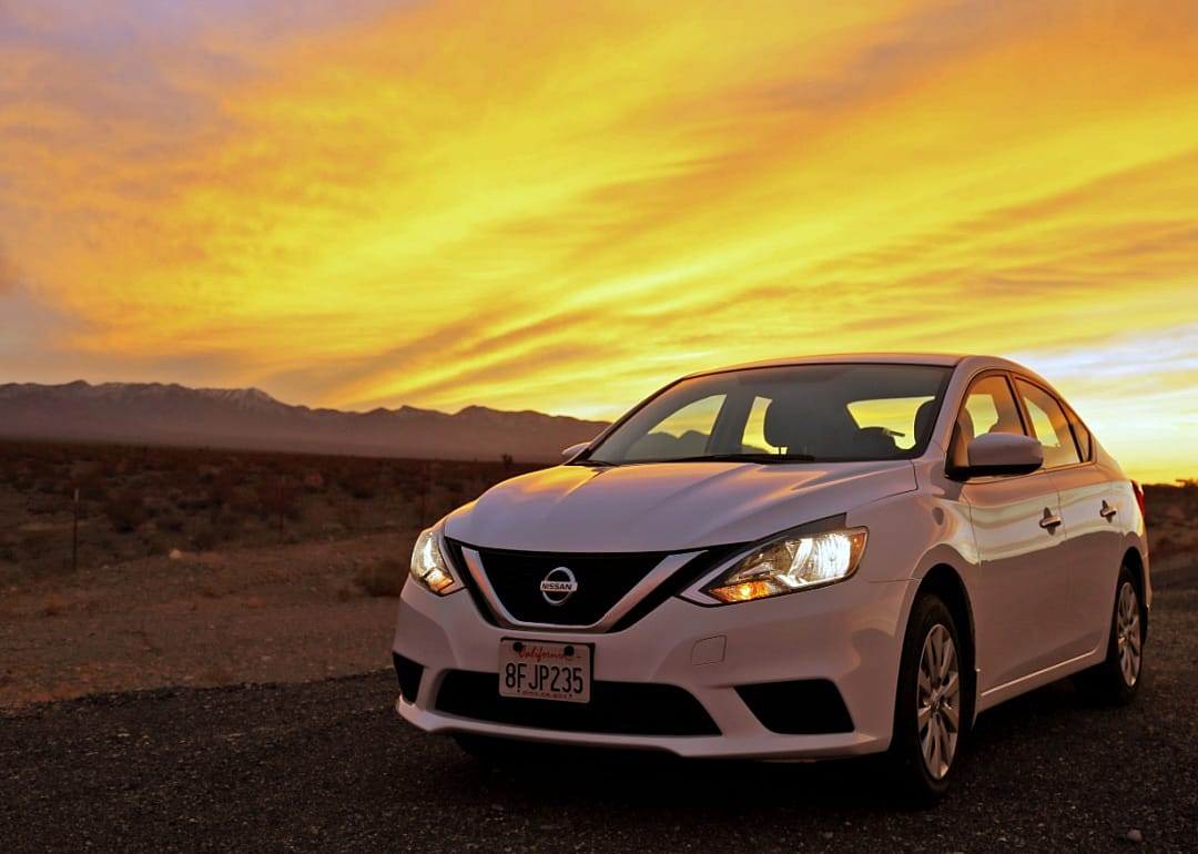 A white Nissan Sentra on a desert road with mountains in the background at sunset.