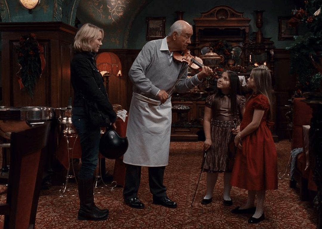 Naomi Watts watching a man in an apron play violin for two little girls in a restaurant.