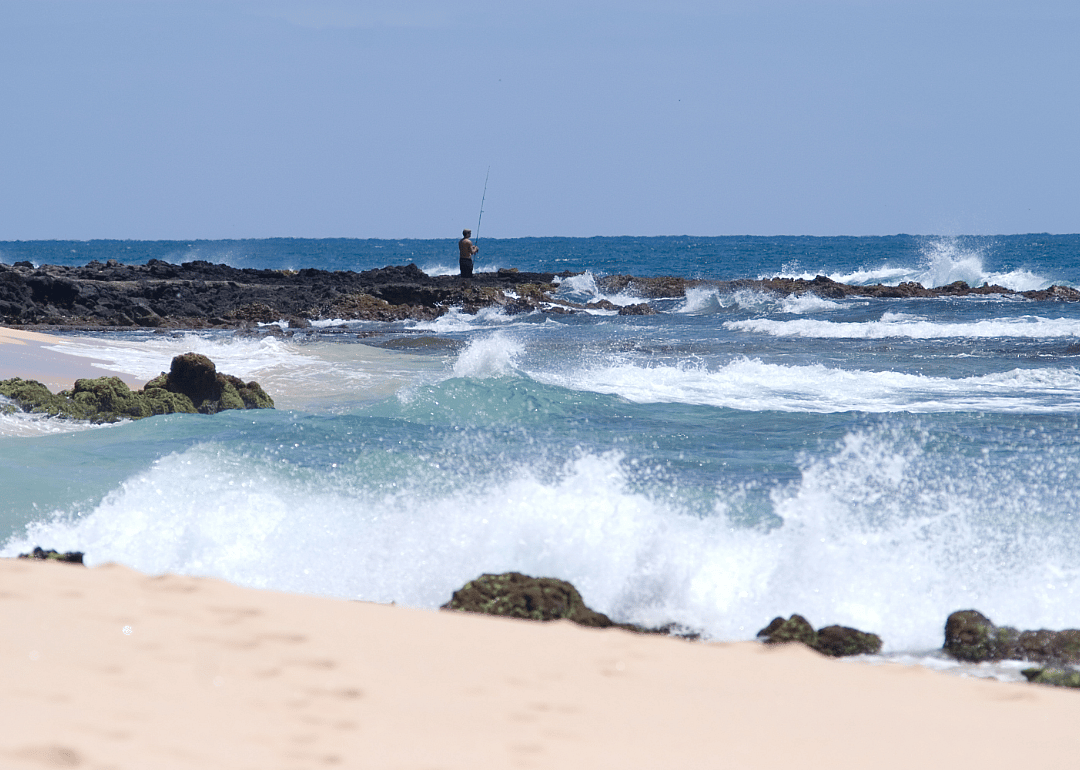 Waves crash onto the rocky, sandy shore while a person fishes in the distance. 