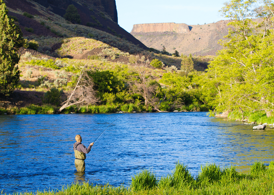 A man in waders fishes in a shallow body of water surrounded by mountains. 