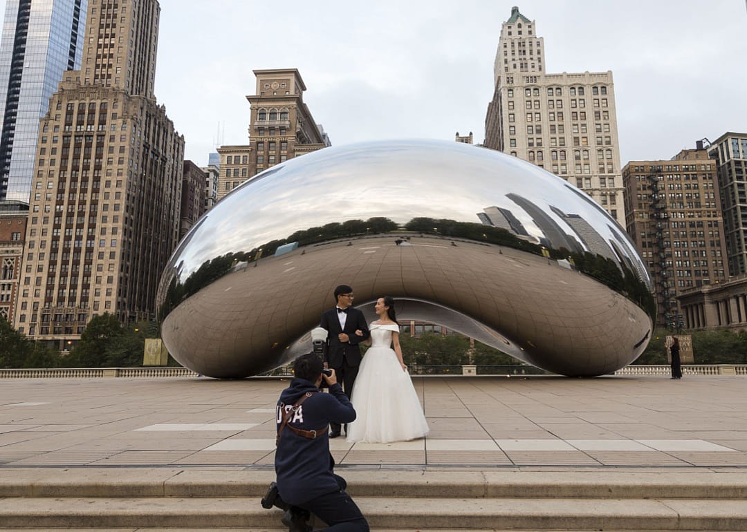 A person photographing a couple in wedding attire in front of a sculpture in downtown Chicago.