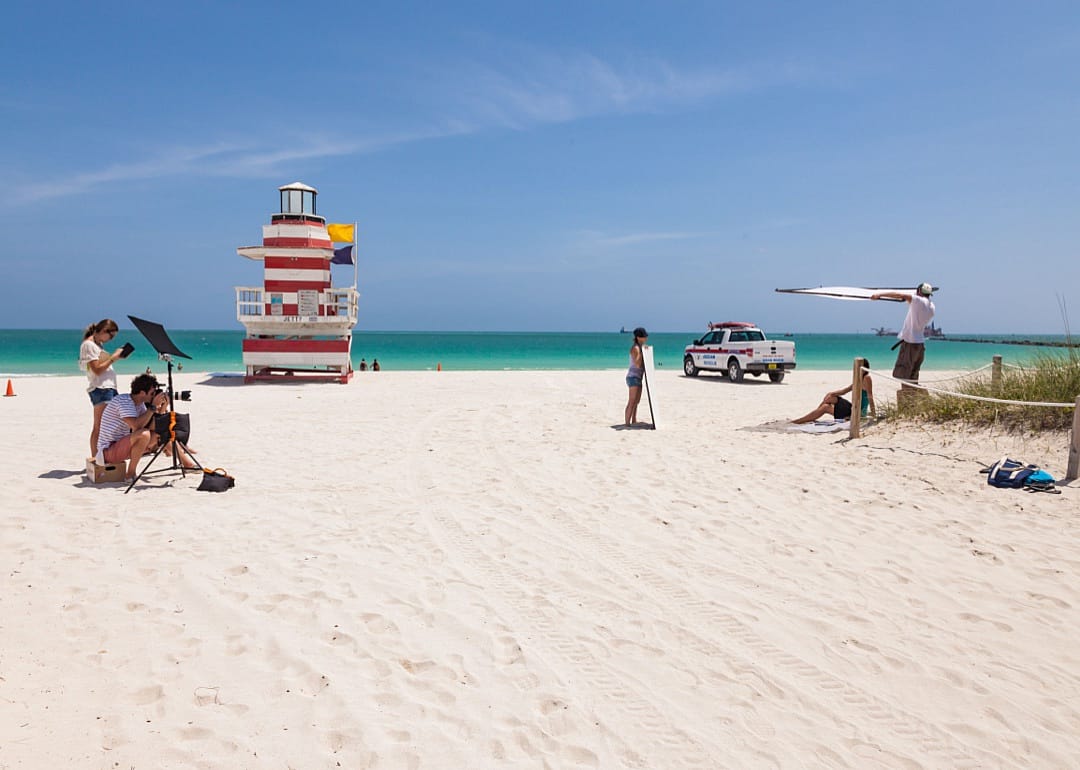 A photographer and their assistants taking photos of someone on the beach with a red and white striped lifeguard stand  by the water.