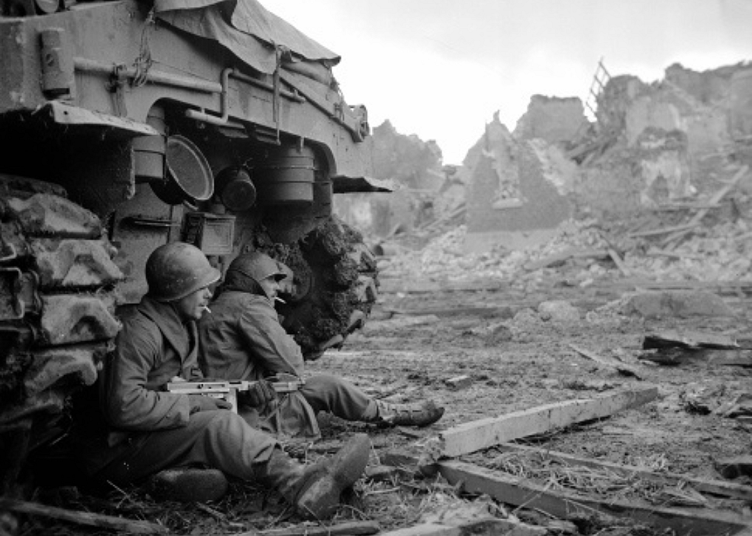 Two men with cigarettes in their mouths and guns in their arms in a war-torn scene seated next to a tank.