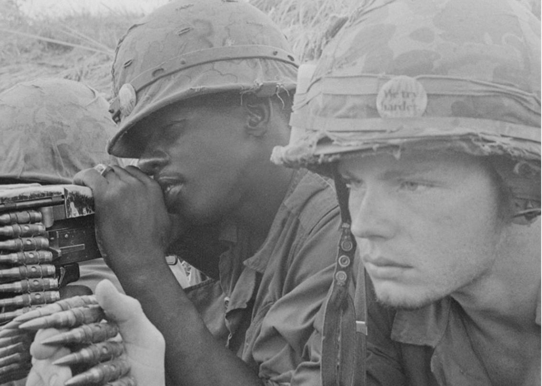 Two soldiers, with pins on their helmets that read, we try harder, holding ammo and a gun.