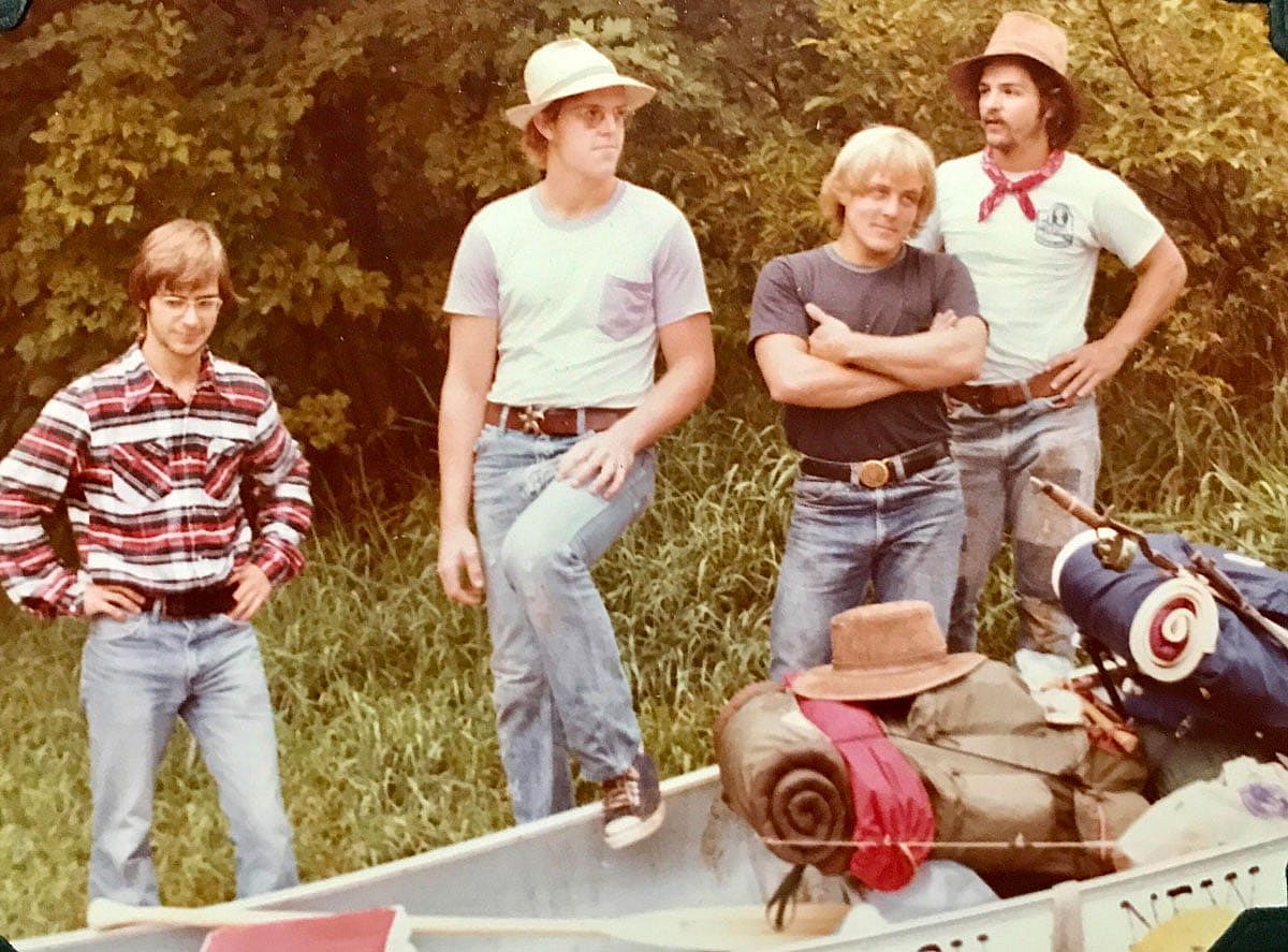 Color photo of four young men standing behind a loaded canoe on a riverbank in 1977. Two wear straw or wide-brimmed hats; one has a red bandana around his neck. The canoe is packed with gear including sleeping rolls and bags. Trees are visible behind them.