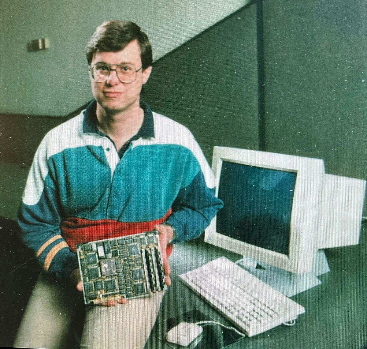 A man in a color-block polo shirt sits next to a desktop computer and monitor, holding a circuit board toward the camera in what appears to be a promotional or magazine photograph from the late 1980s.