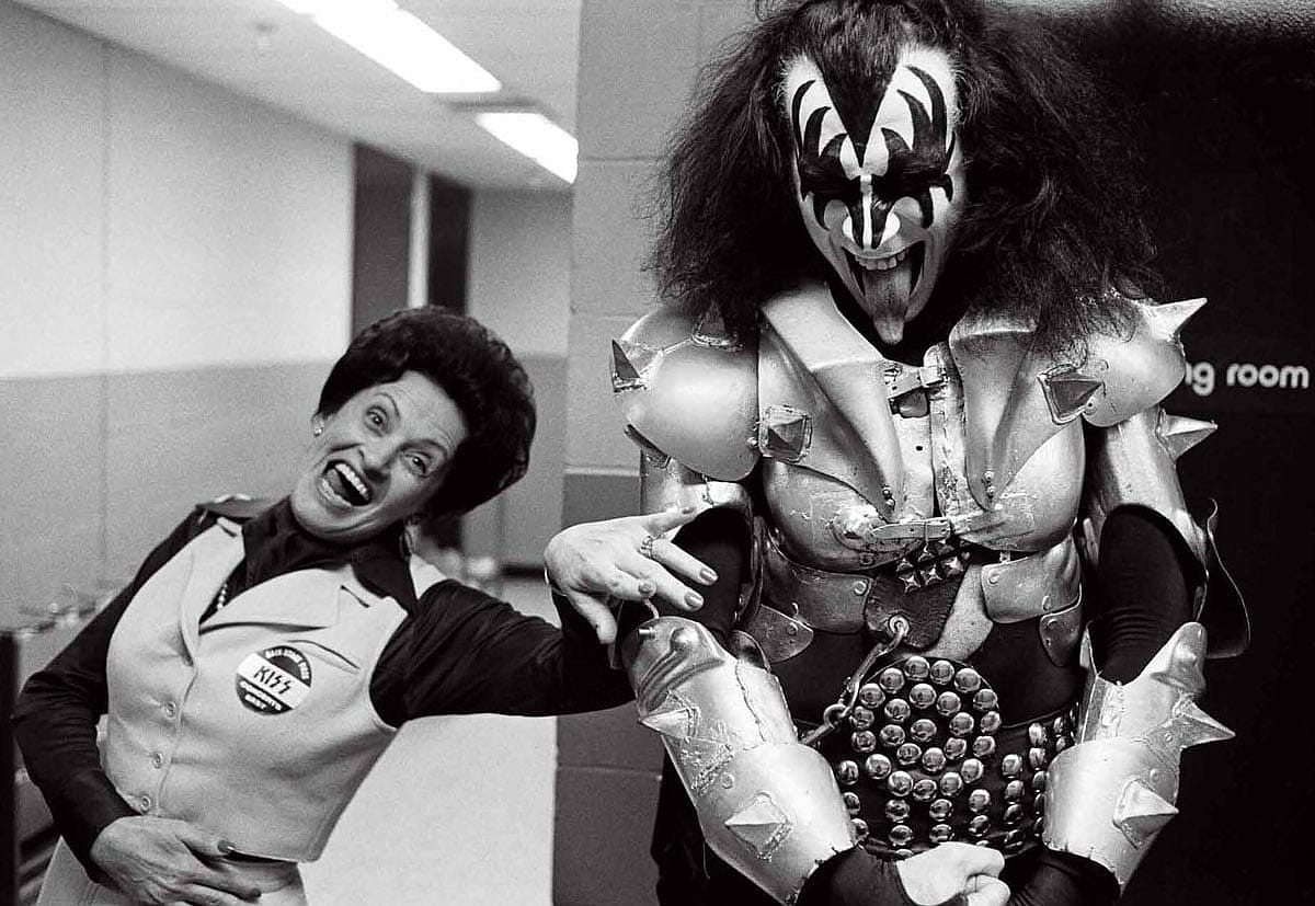 Black-and-white photo of Gene Simmons in full KISS Demon costume and makeup standing backstage with his mother Florence in 1977. Florence wears a vest with a KISS button pinned to it and laughs openly at the camera. Gene, in spiked armor and black and white face paint, stands beside her grinning.