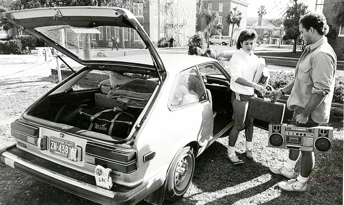 Black and white photo of a woman unpacking her car and handing a case of cassettes to a man holding a boom box