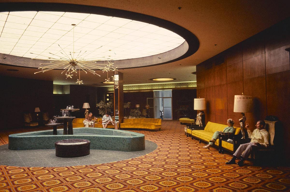 Color photo of the ornate lobby of the Nevele Hotel in Ellenville, New York, in 1977. A large oval skylight illuminates the space from above, beneath which hang Sputnik-style chandeliers. A kidney-shaped turquoise fountain sits in the center of a boldly patterned red and gold carpet. Several guests sit on gold velvet sofas along the walls, and a small indoor fountain is visible in the background.