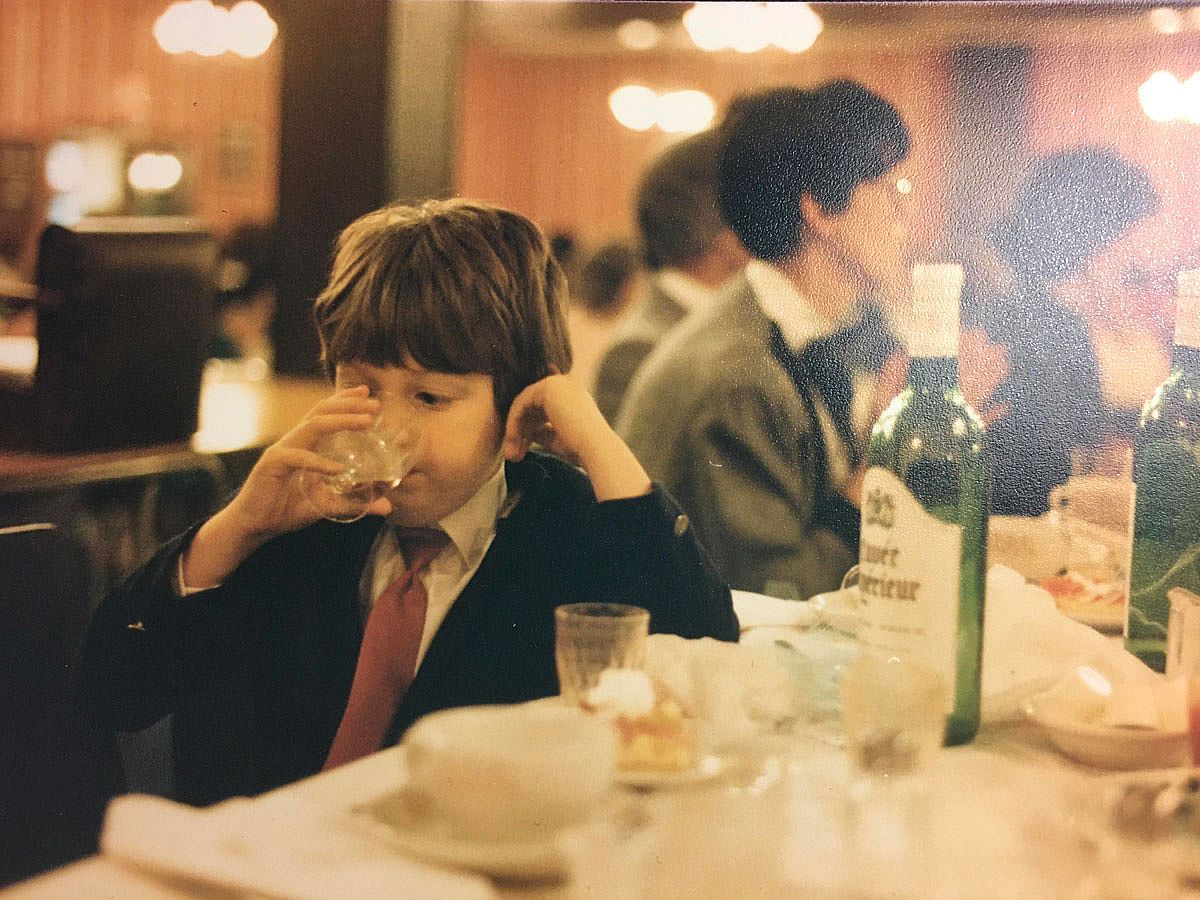A young boy in a suit and tie sips from a wine glass at a restaurant table, with a wine bottle and other glasses visible nearby and adults seated behind him.