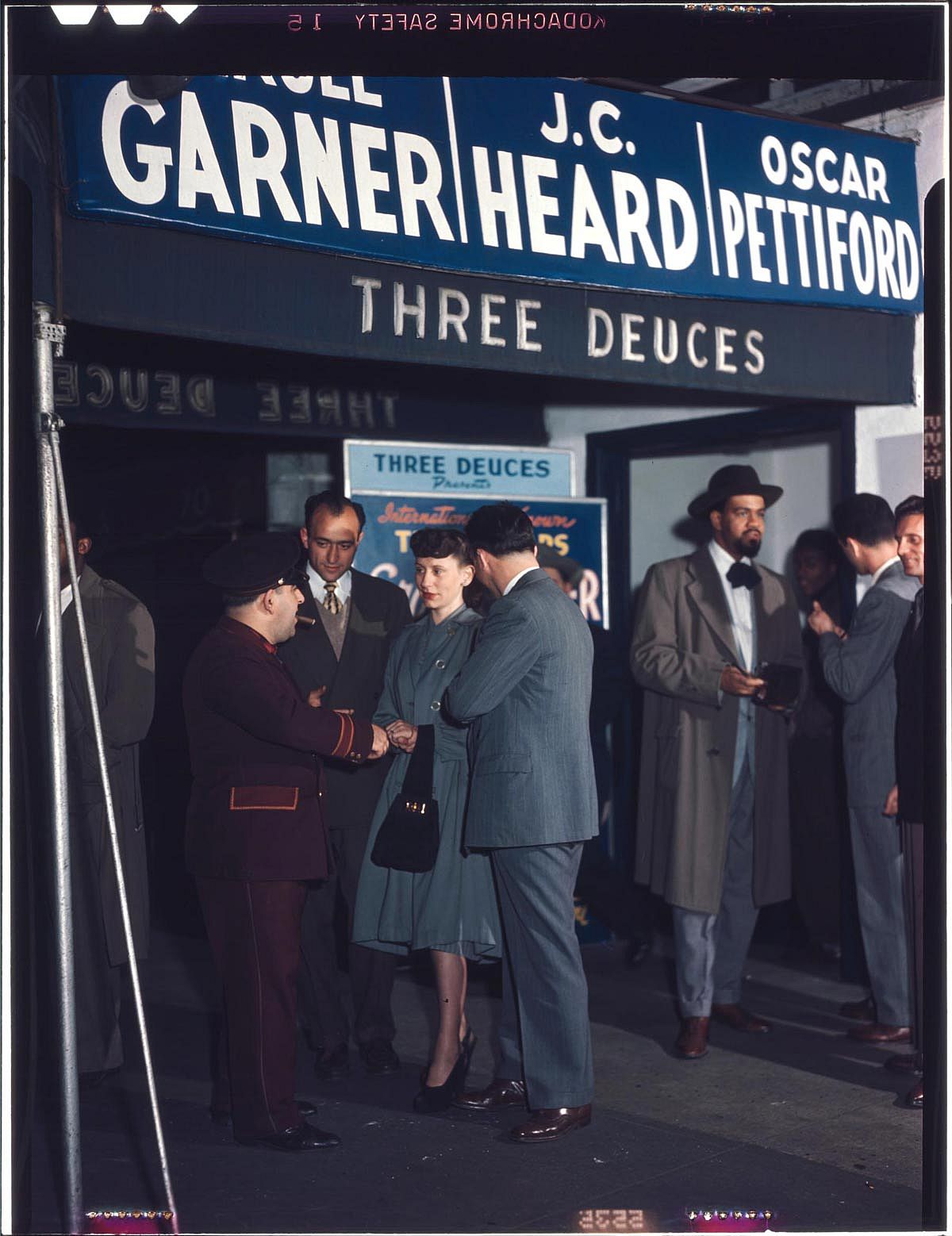 Color photograph of well-dressed patrons standing outside The Three Deuces nightclub entrance on 52nd Street in New York City, circa 1948, beneath a large blue marquee displaying the names of jazz performers Erroll Garner, J.C. Heard, and Oscar Pettiford