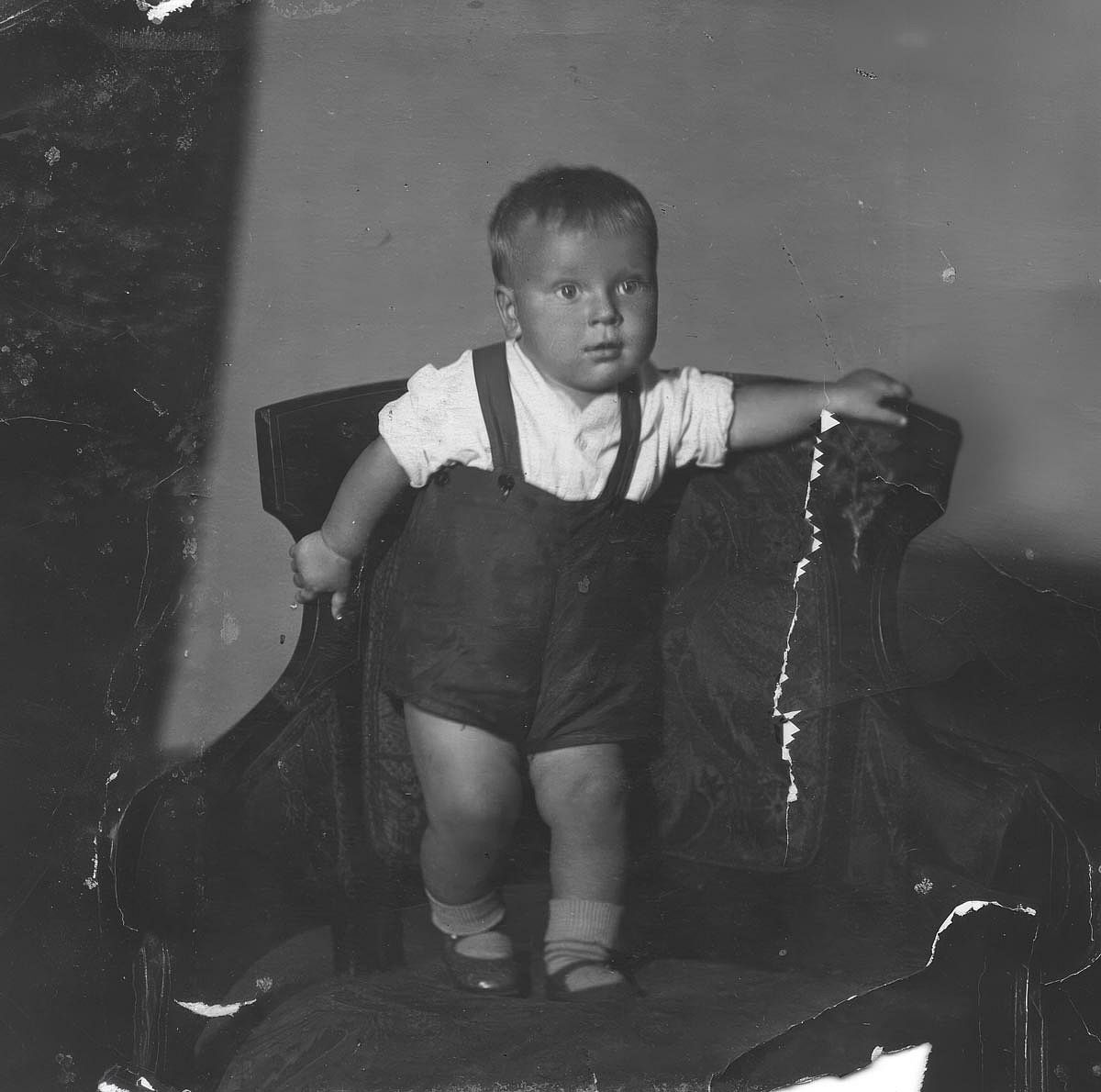 Baby boy in white shirt and dark suspender shorts standing on upholstered armchair, scanned archival black and white print circa 1940s