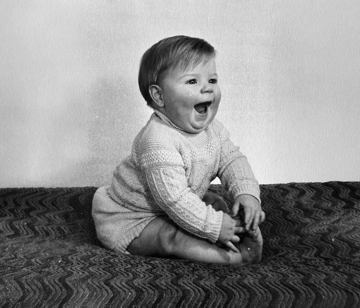 Black and white photo of a baby boy in the 1940s sitting on a bed.