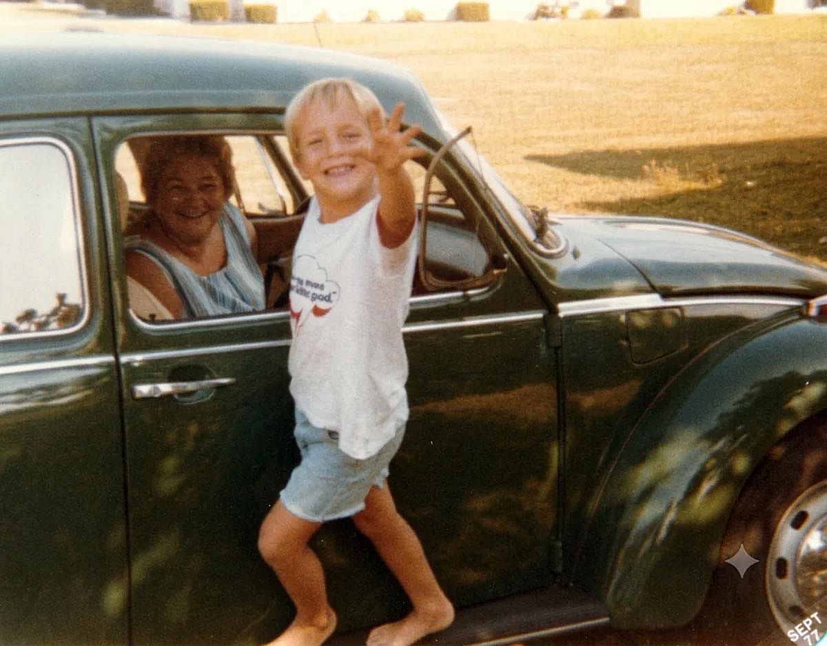 Smiling kid hanging off the side of a green VW Bug while his mom is sitting behind the drivers wheel.